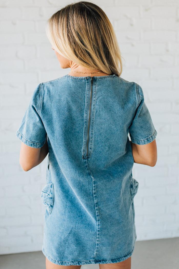 Blonde woman wearing a denim shift dress to the knee with short sleeves, side pickets and bow accents on each pocket.
