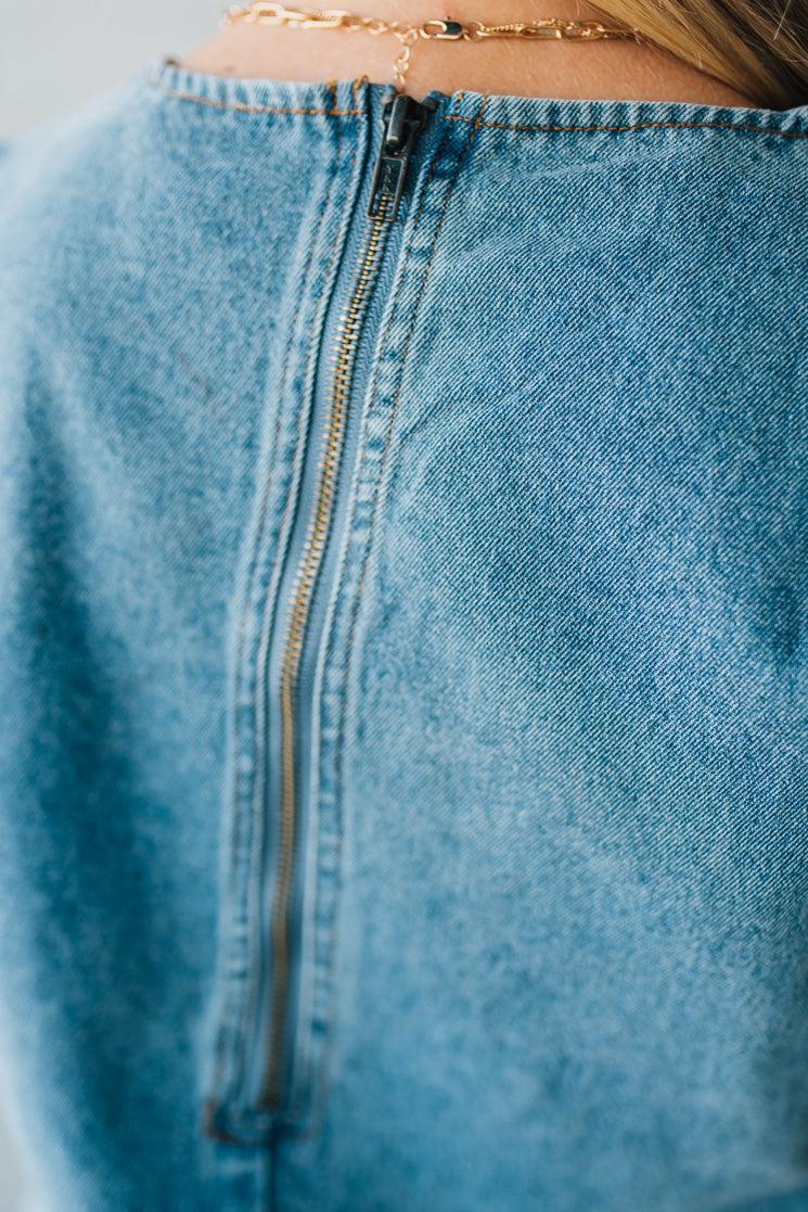 Blonde woman wearing a denim shift dress to the knee with short sleeves, side pickets and bow accents on each pocket.