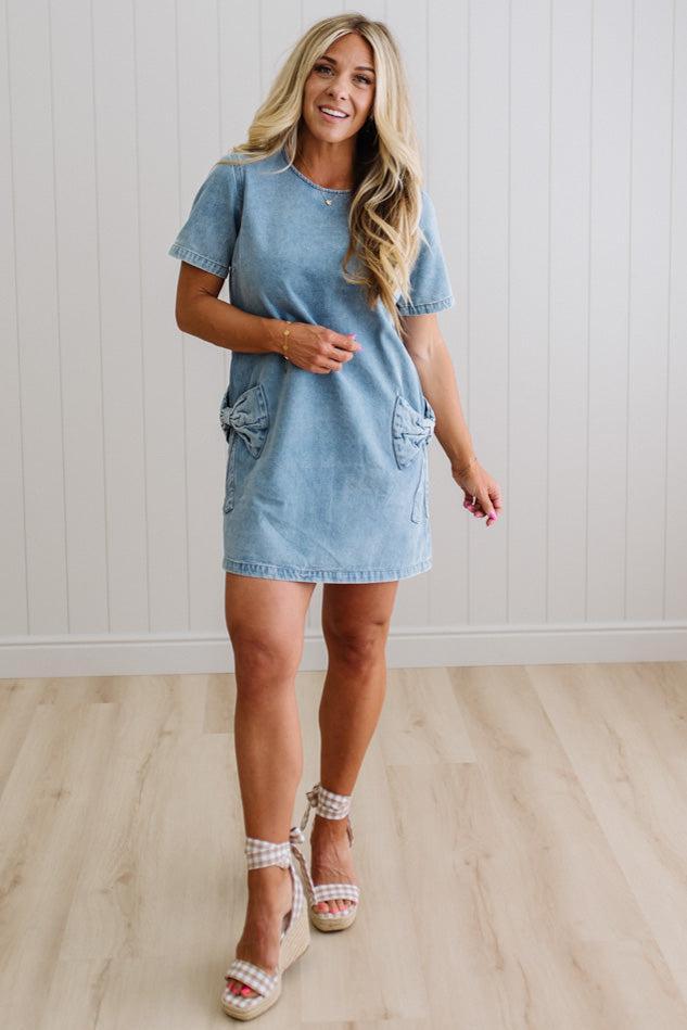 Blonde woman wearing a denim shift dress to the knee with short sleeves, side pickets and bow accents on each pocket.