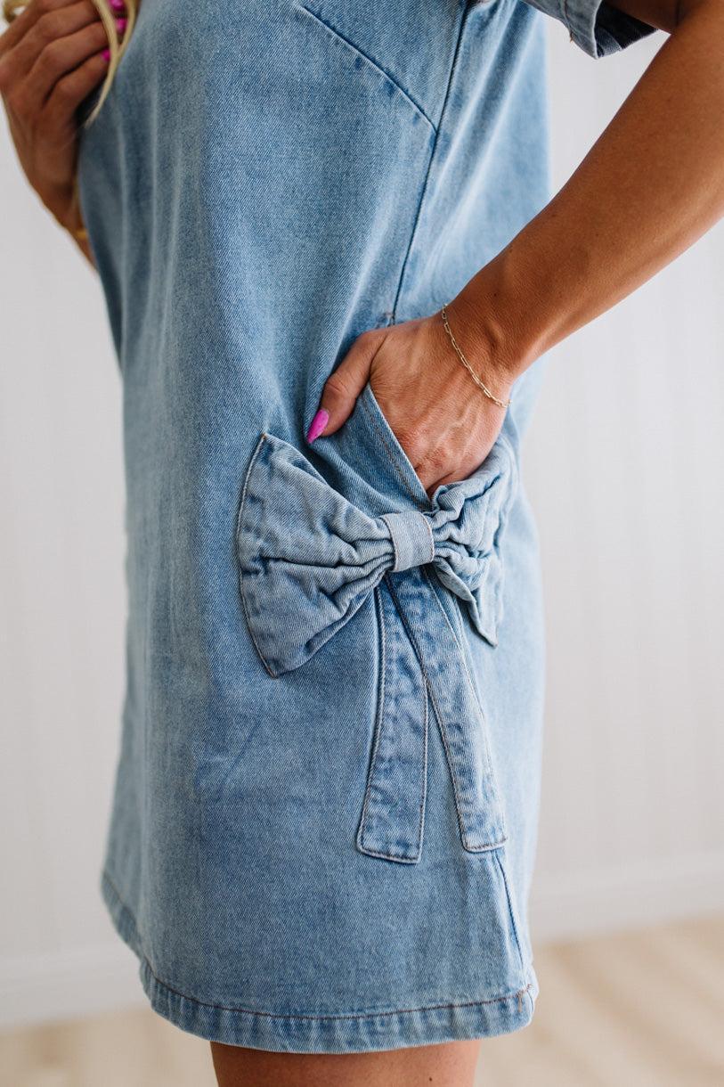 Blonde woman wearing a denim shift dress to the knee with short sleeves, side pickets and bow accents on each pocket.