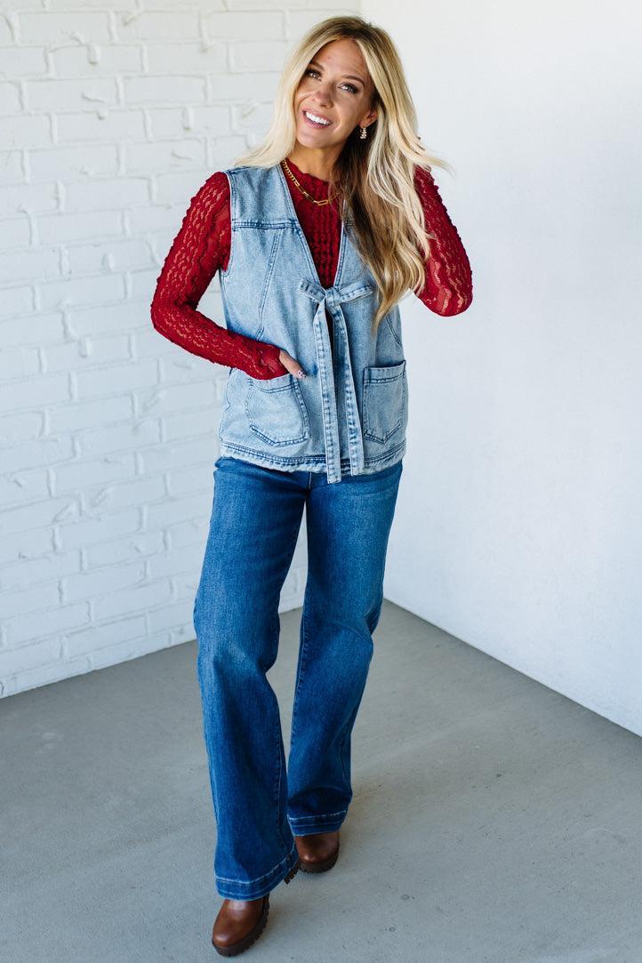 Woman wearing a denim vest over a red top with blue jeans against a white brick wall.