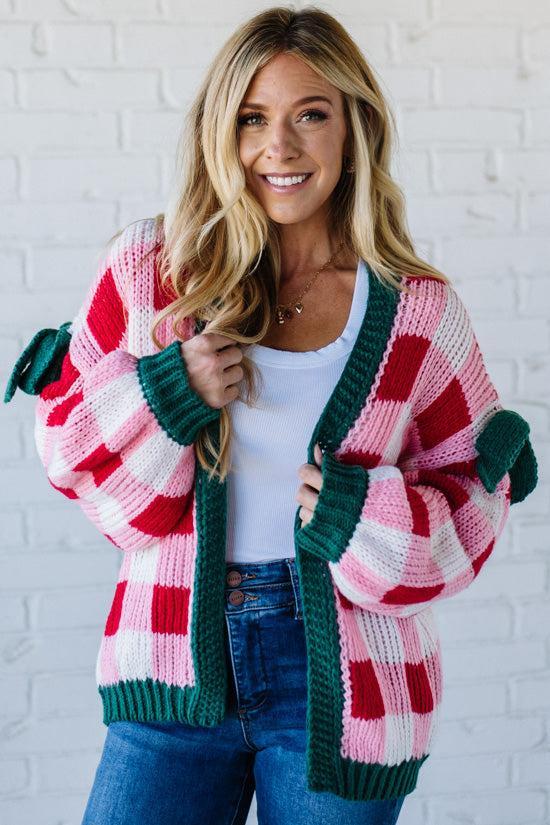Woman wearing a colorful plaid cardigan with pom-pom details against a white brick wall.