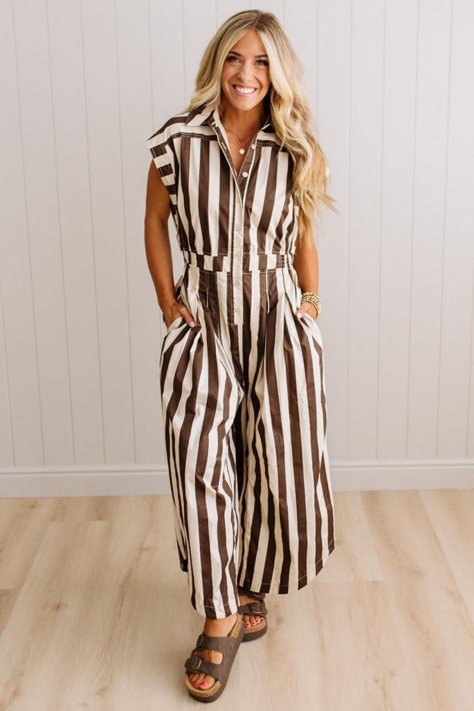 BLONDE WOMAN WEARING A SLEEVELESS WIDE LEG, WIDE STRIPE JUMPSUIT STANDING ON A LIGHT WOOD FLOOR AND NEUTRAL WALL BEHIND.