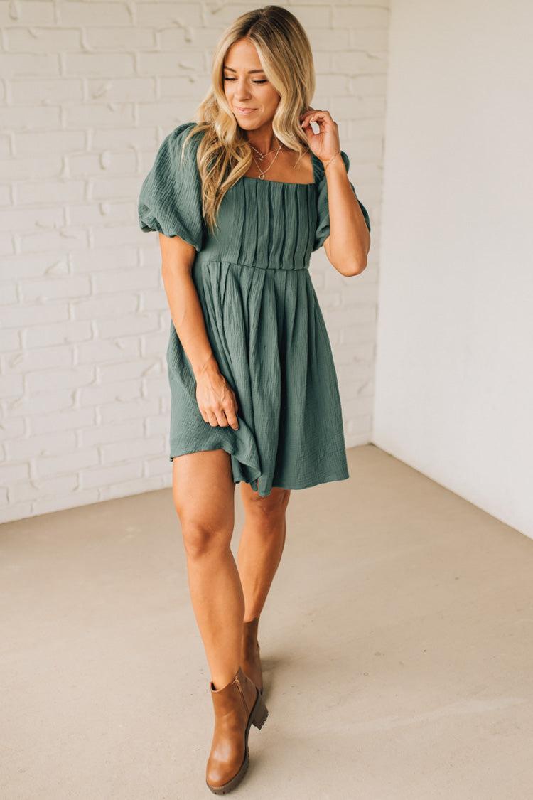 Woman wearing a green dress with short sleeves against a white brick wall.