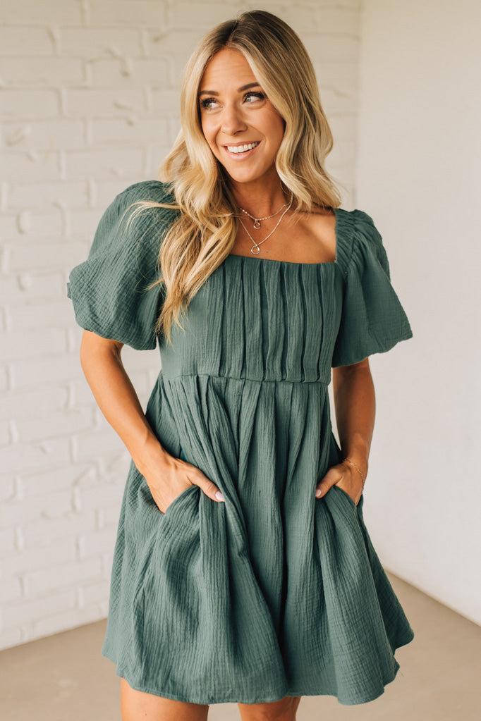 Woman wearing a green dress with puff sleeves against a white brick wall.