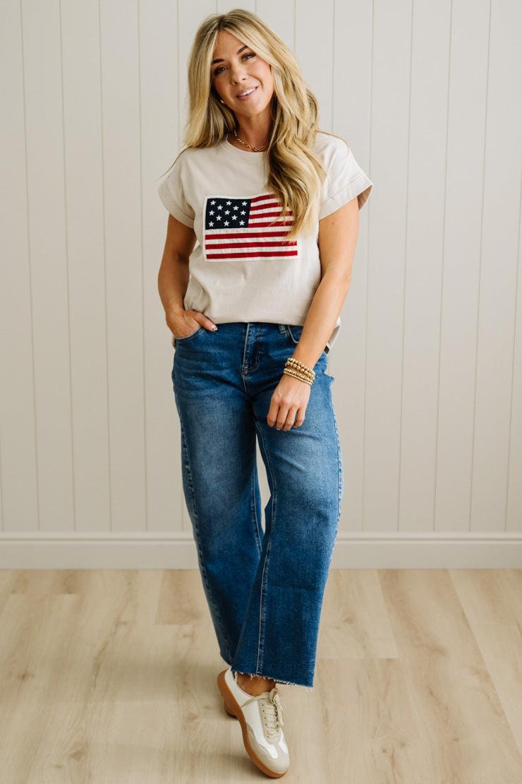 Woman wearing a t-shirt with an American flag design and blue jeans against a white wall.