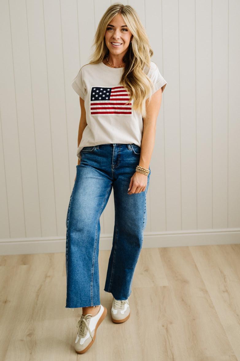 Woman wearing a white t-shirt with an American flag design and blue jeans, standing against a light wooden paneled wall.