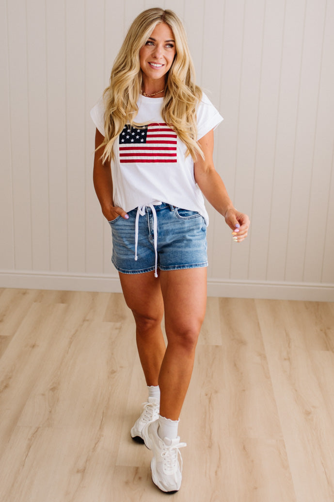Woman wearing a white t-shirt with an American flag design and denim shorts on a wooden floor.