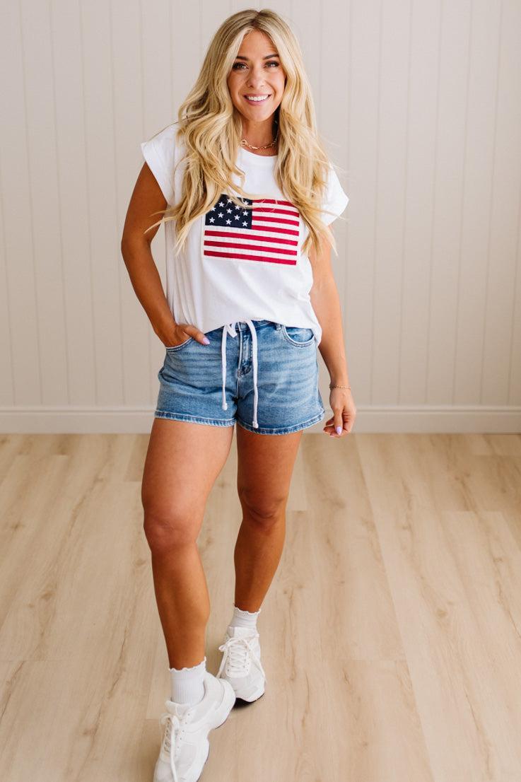 Woman wearing a white t-shirt with an American flag design and denim shorts on a wooden floor.