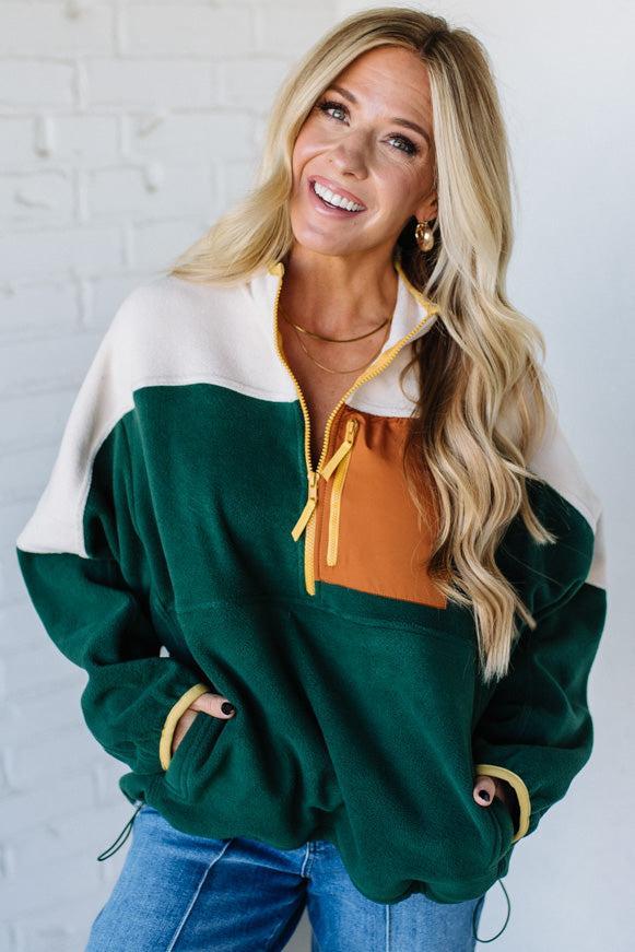 Woman wearing a green, white, and brown color-blocked jacket against a white brick wall.