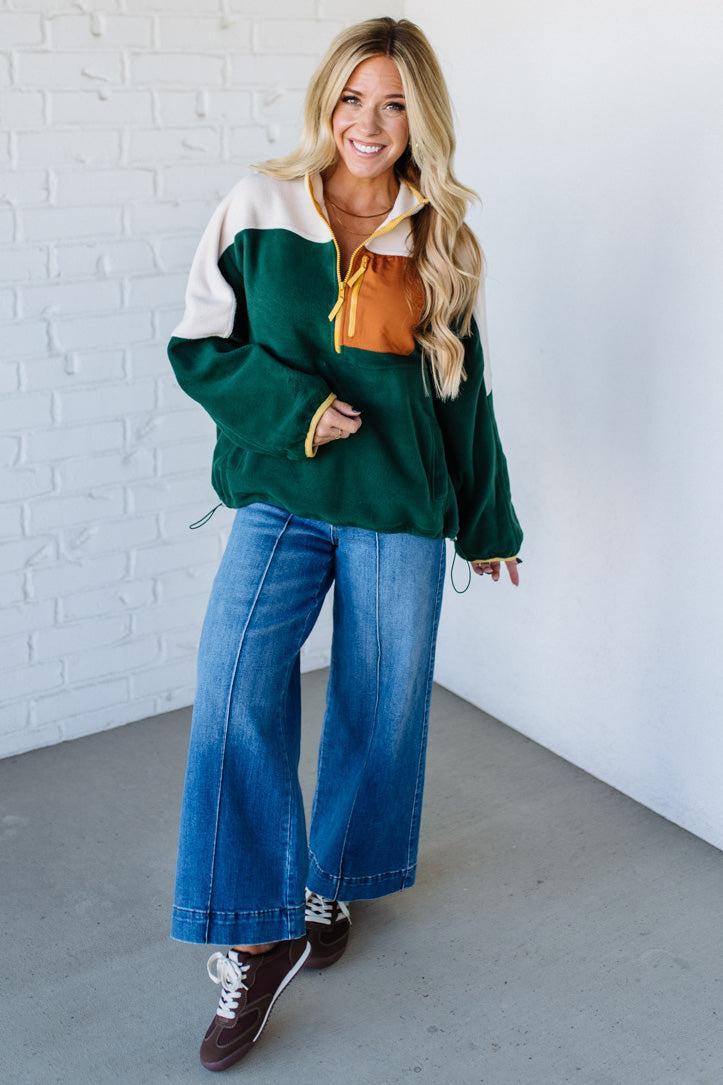 Woman wearing a green and white jacket with a mustard shirt, blue jeans, and brown shoes against a white brick wall.