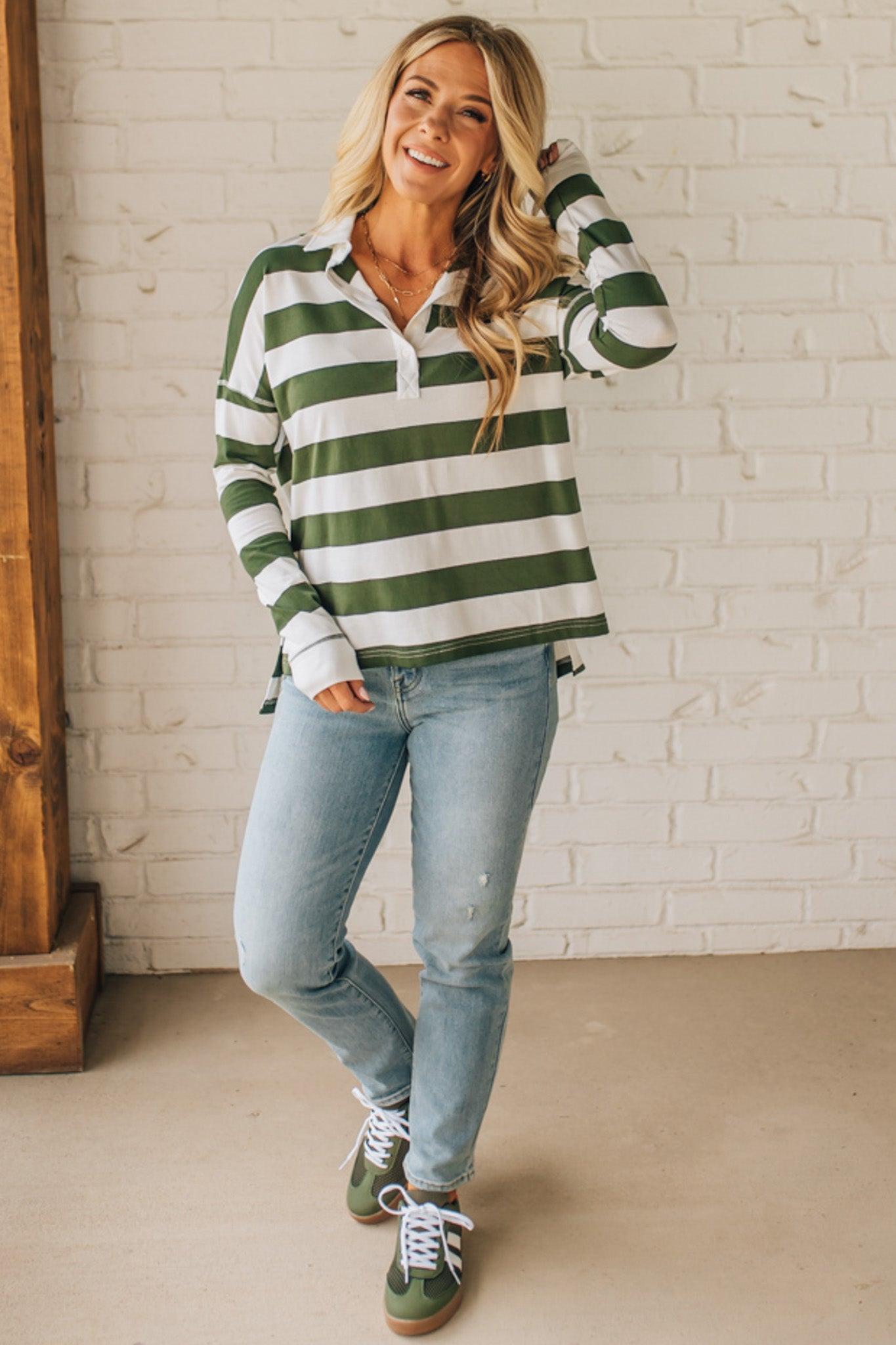 Woman wearing a green and white striped sweater with blue jeans against a white brick wall.