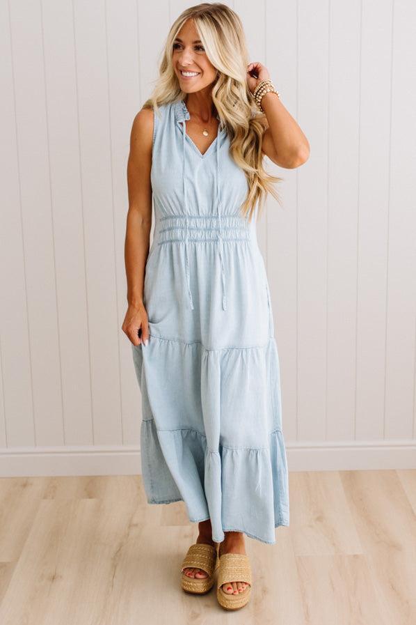 A woman modeling a sleeveless light blue tiered maxi dress with a smocked waist and V-neckline, paired with tan slide sandals, standing against a white paneled wall and light wood floor.