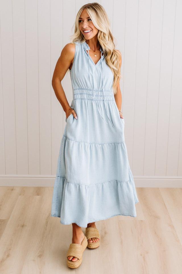A woman modeling a sleeveless light blue tiered maxi dress with a smocked waist and side pockets, paired with beige slide sandals, standing in front of a white paneled wall and light wood floor.