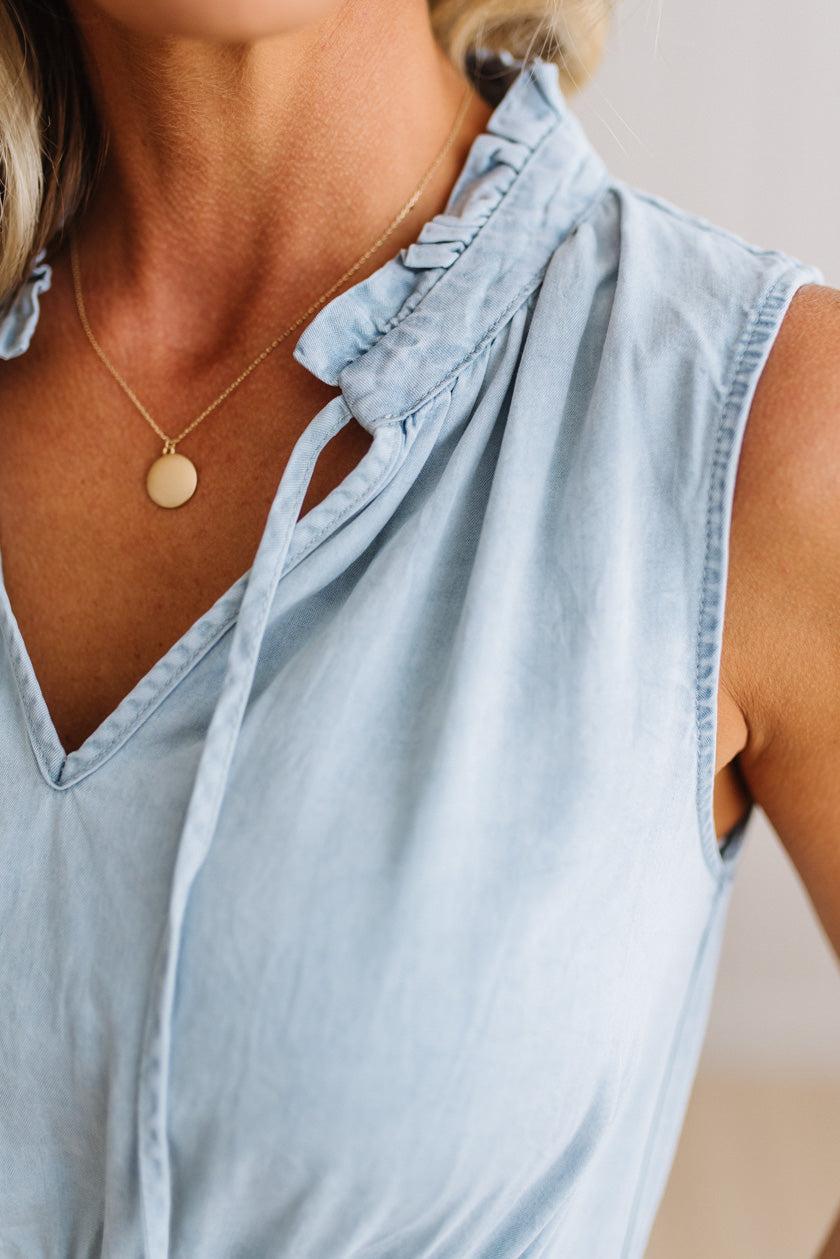 A close-up of a woman wearing a light blue sleeveless top with a ruffled collar and tie detail, paired with a delicate gold pendant necklace.