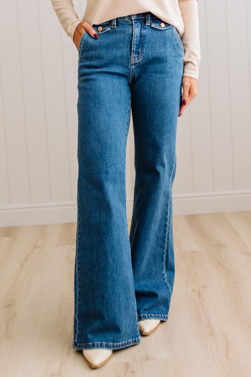 WOMAN WEARING HIGH RISE FLARE JEANS IN A DARK WASH AND BUTTON FLAP SMALL POCKETS AT THE HIP.