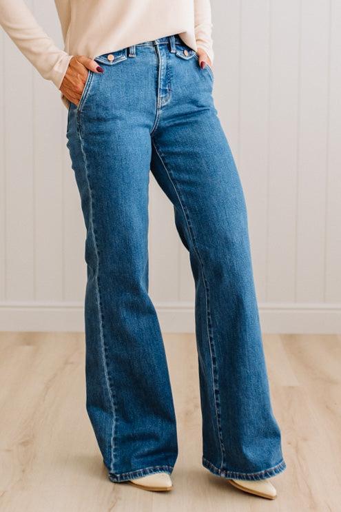 WOMAN WEARING HIGH RISE FLARE JEANS IN A DARK WASH AND BUTTON FLAP SMALL POCKETS AT THE HIP.
