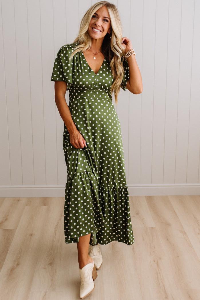 A woman modeling a green midi dress with white polka dots, featuring short flutter sleeves, a V-neckline, and a tiered skirt, paired with white cowboy boots against a light wood-paneled wall.