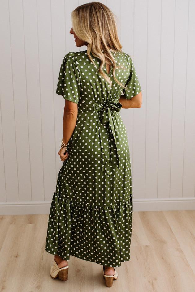 A woman wearing a green and white polka dot midi dress with short puff sleeves and a tie waist, shown from the back against a white paneled wall and light wood floor.
