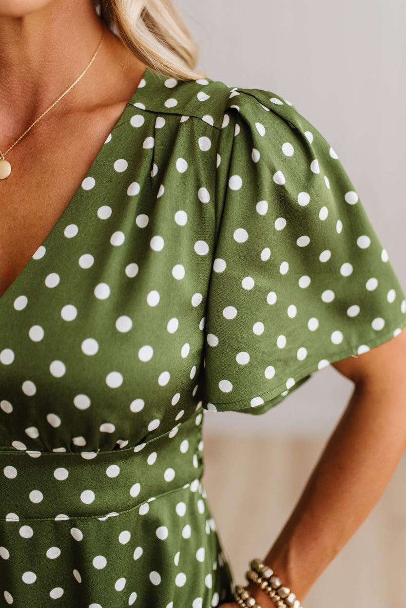 Close-up of a woman wearing an olive green wrap-style dress with white polka dots, short puff sleeves, and a V-neckline.