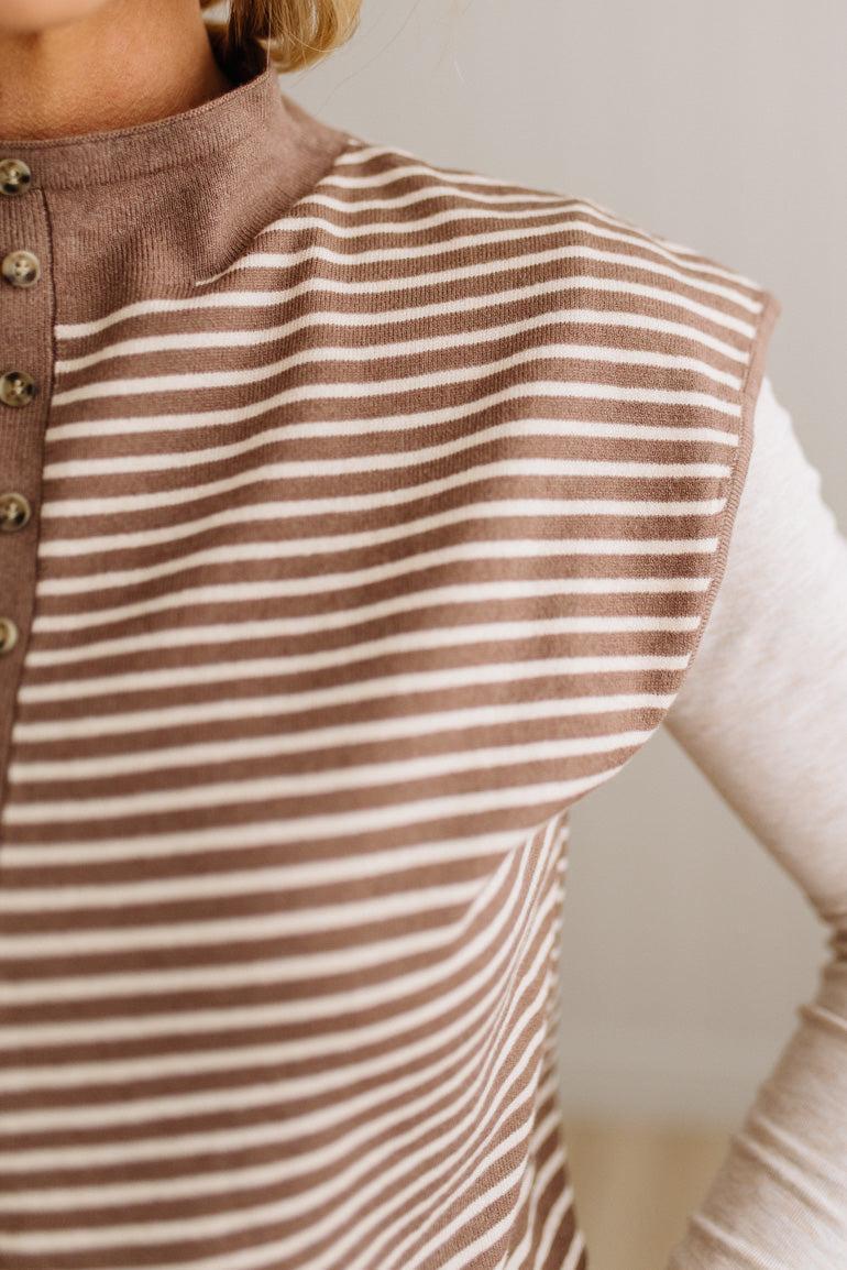BLONDE WOMAN WEARING A MOCK NECK, FRONT BUTTON DETAILED, WITH CAP SLEEVES AND STRIPES. STANDING IN A WELL LIT STUDIO.
