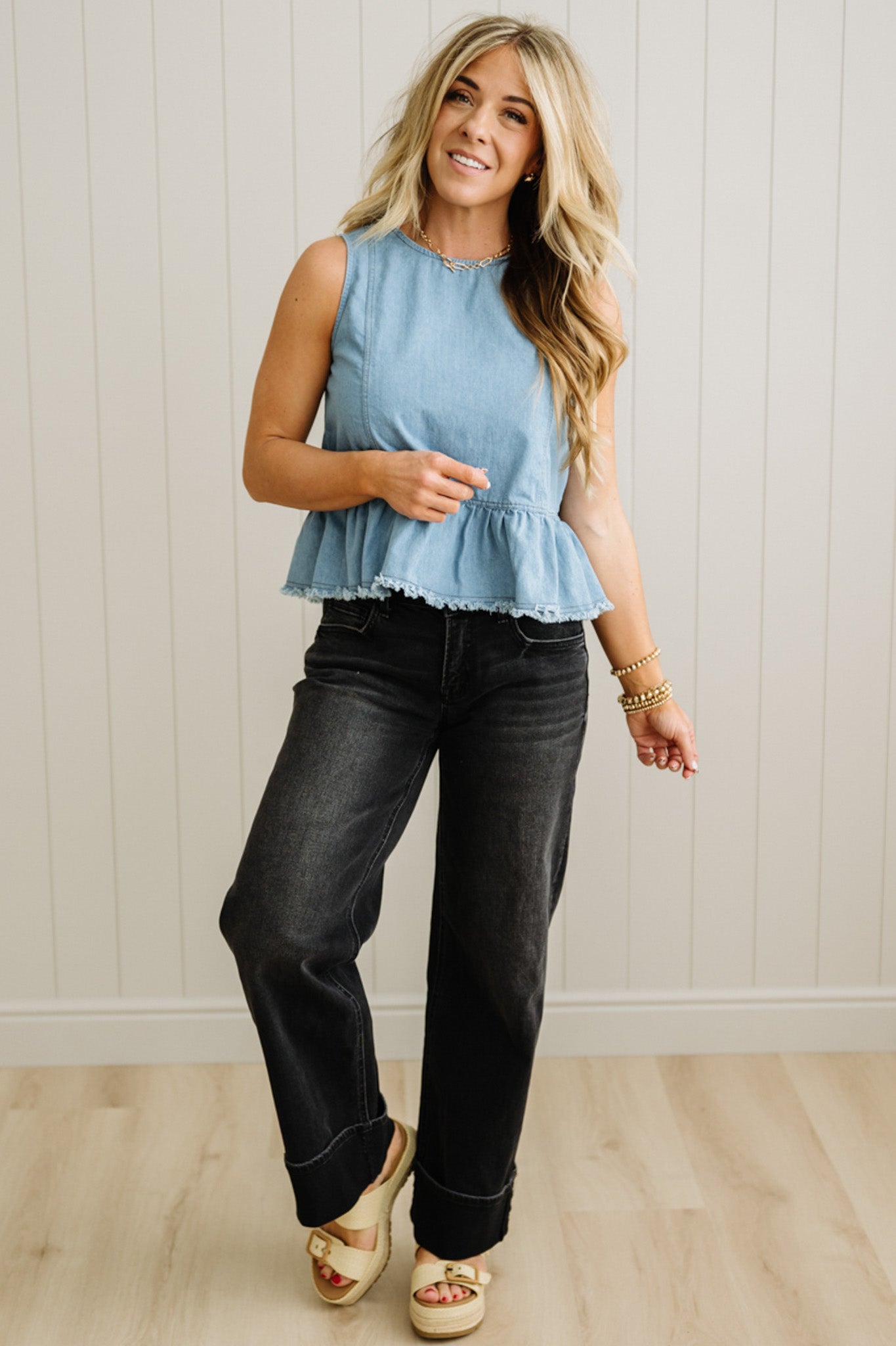 Woman wearing a light blue sleeveless top and black jeans against a white paneled wall.