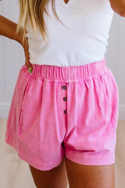 Person wearing pink shorts with buttons and a white top on a neutral background