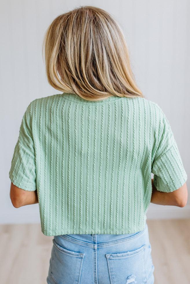 A woman viewed from behind wearing a mint green short-sleeve ribbed knit top and light blue jeans, standing against a plain light gray wall.
