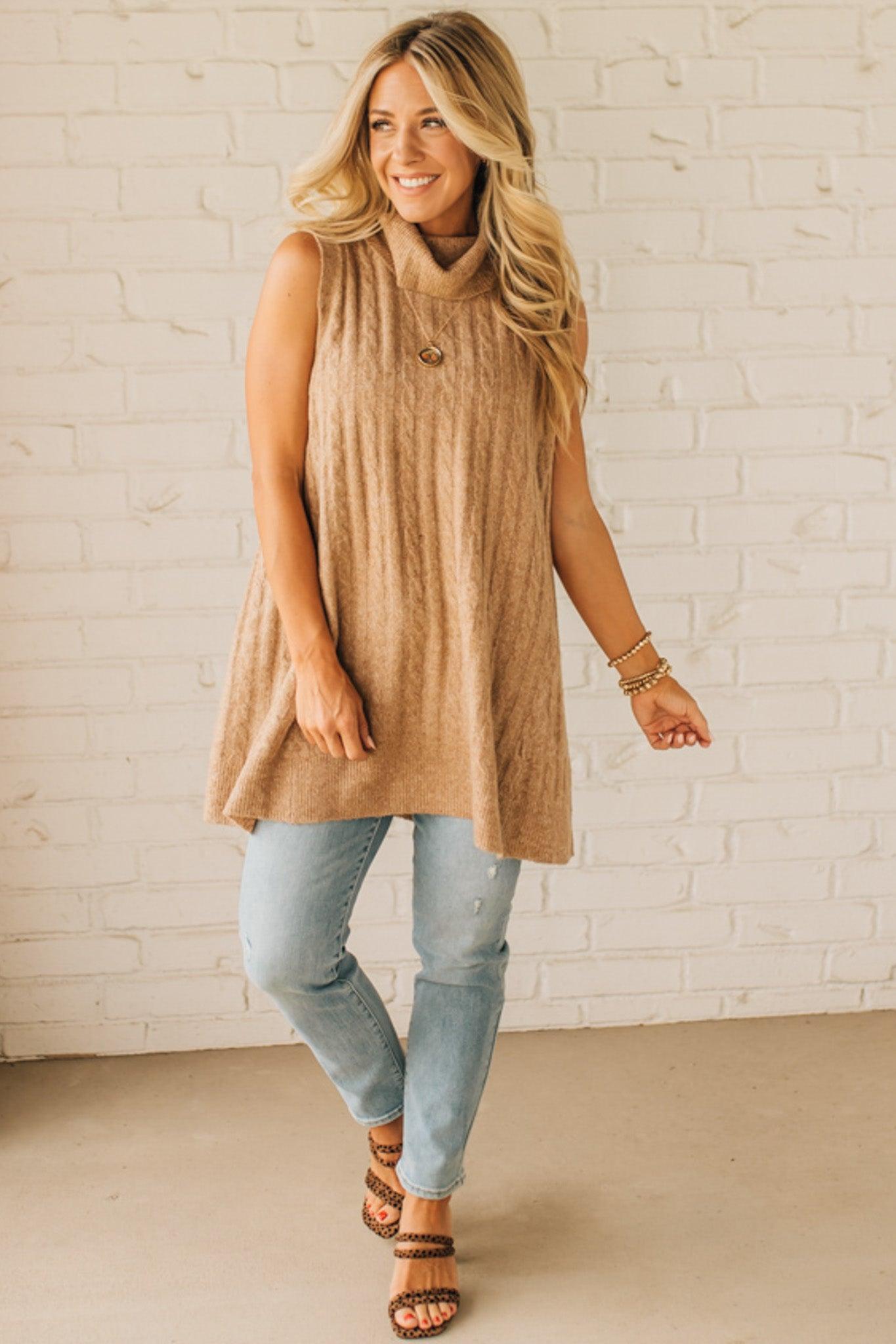 Woman wearing a beige sleeveless dress and light blue jeans against a white brick wall.