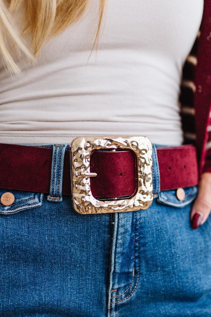 A rippled square frame buckle with a molten, hammered gold-tone finish sits front and center, on a burgundy faux suede belt.