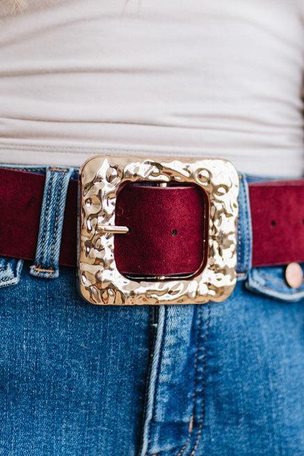 A rippled square frame buckle with a molten, hammered gold-tone finish sits front and center, on a burgundy faux suede belt.