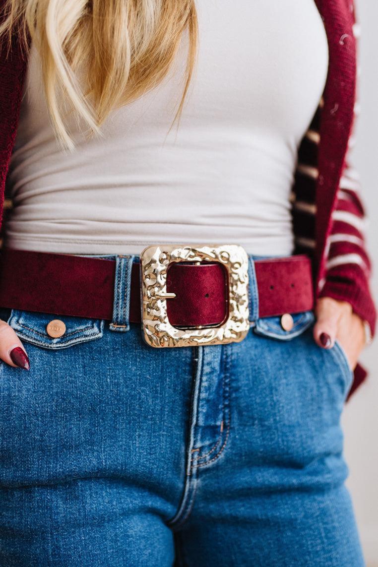 A rippled square frame buckle with a molten, hammered gold-tone finish sits front and center, on a burgundy faux suede belt.
