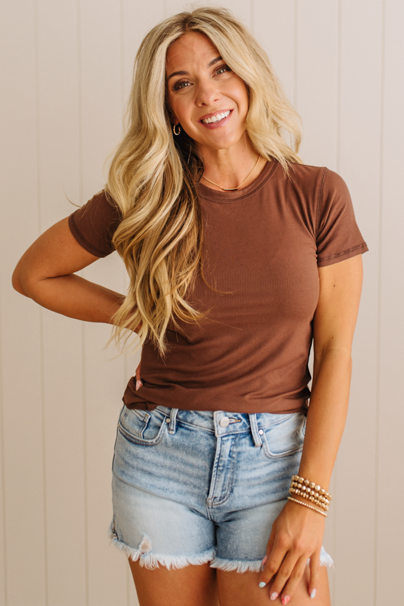 Woman wearing a brown t-shirt and denim shorts against a beige wall.