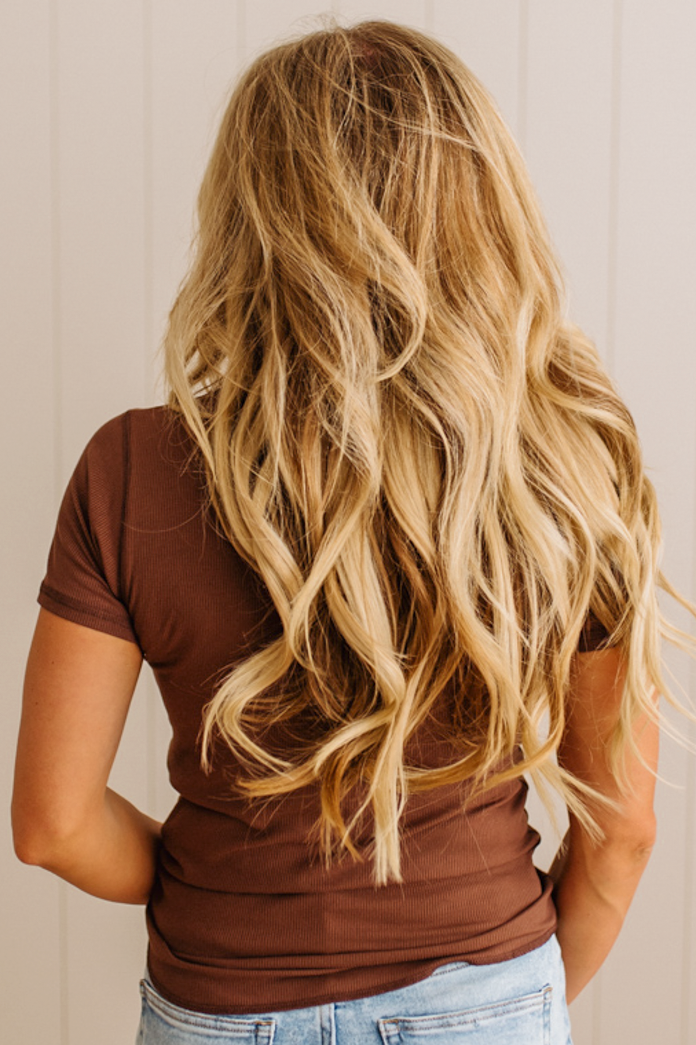 Person with long blonde hair wearing a brown shirt and blue jeans against a neutral background