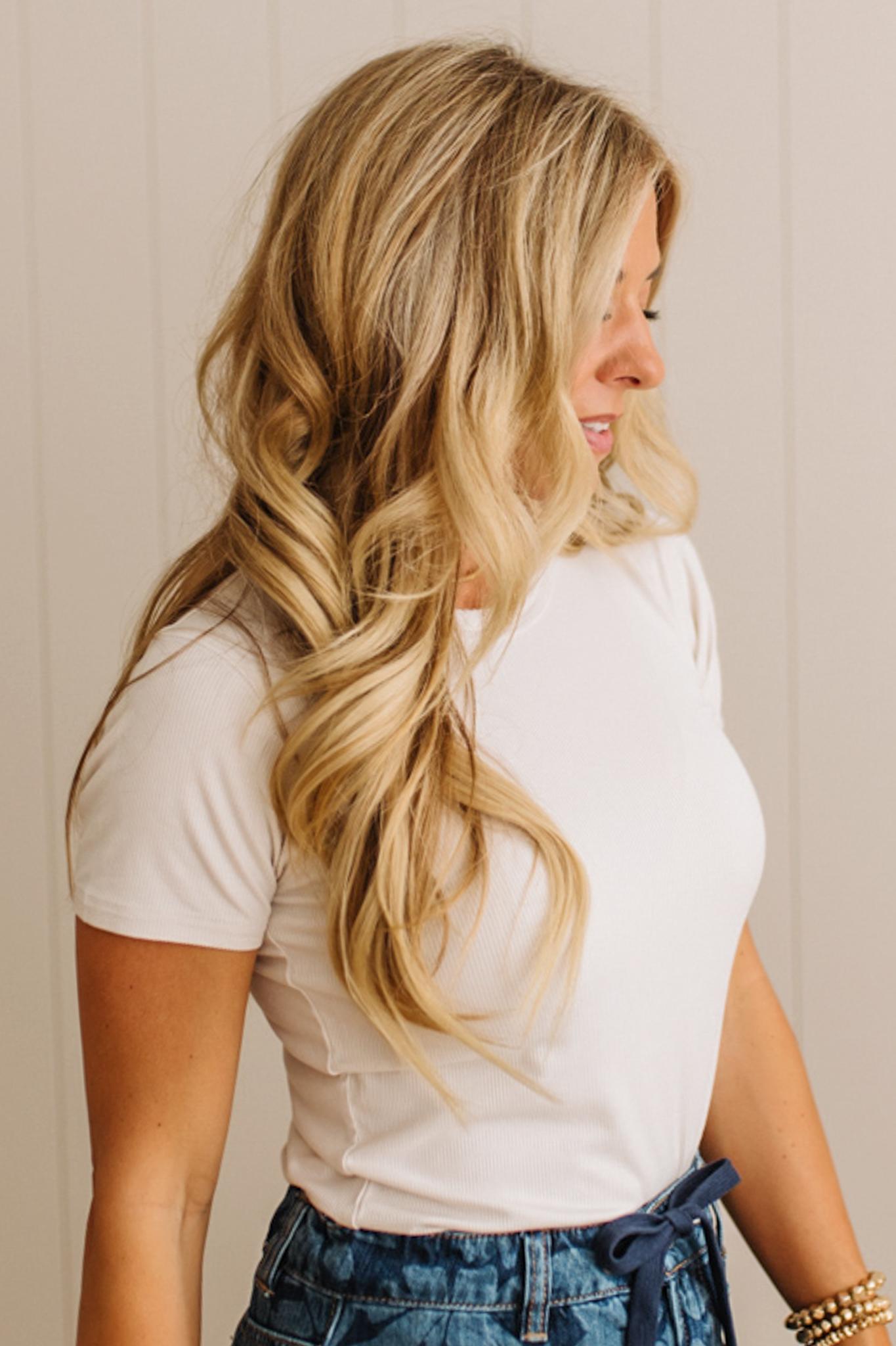 Woman with long blonde hair wearing a white top and blue jeans against a neutral background