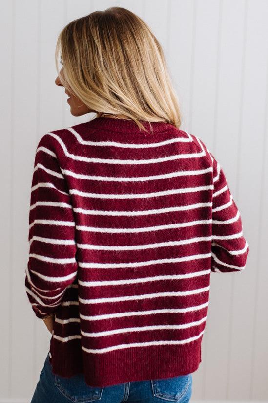 BLONDE WOMAN IN A BRIGHTLY LIT STUDIO WEARING A CREAM TANK AND DARK JEANS SHOWING A OUTSEAMED, LONG SLEEVE, FUZZY, CREW NECK STRIPED BUTTON DOWN SWEATER CARDIGAN IN BURGUNDY.
