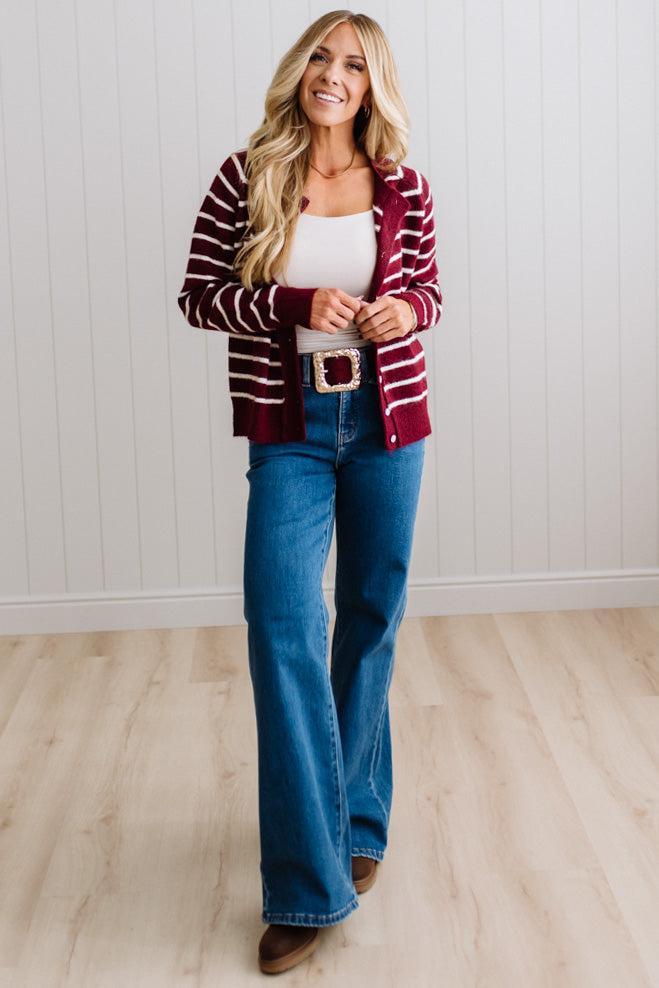 BLONDE WOMAN IN A BRIGHTLY LIT STUDIO WEARING A CREAM TANK AND DARK JEANS SHOWING A OUTSEAMED, LONG SLEEVE, FUZZY, CREW NECK STRIPED BUTTON DOWN SWEATER CARDIGAN IN BURGUNDY.