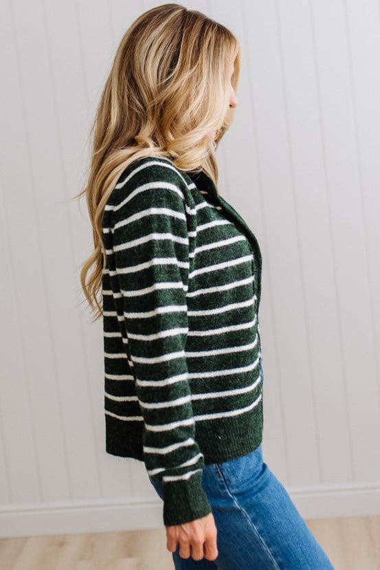 BLONDE WOMAN IN A BRIGHTLY LIT STUDIO WEARING A CREAM TANK AND DARK JEANS SHOWING A OUTSEAMED, LONG SLEEVE, FUZZY, CREW NECK STRIPED BUTTON DOWN SWEATER CARDIGAN IN FOREST GREEN.
