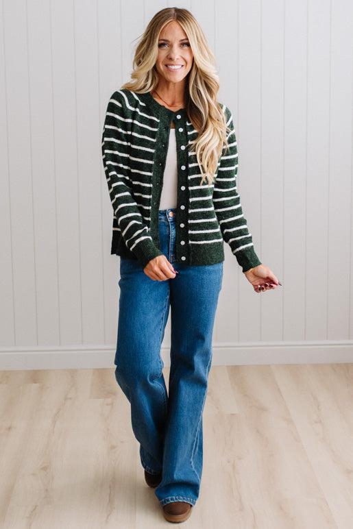 BLONDE WOMAN IN A BRIGHTLY LIT STUDIO WEARING A CREAM TANK AND DARK JEANS SHOWING A OUTSEAMED, LONG SLEEVE, FUZZY, CREW NECK STRIPED BUTTON DOWN SWEATER CARDIGAN IN FOREST GREEN.