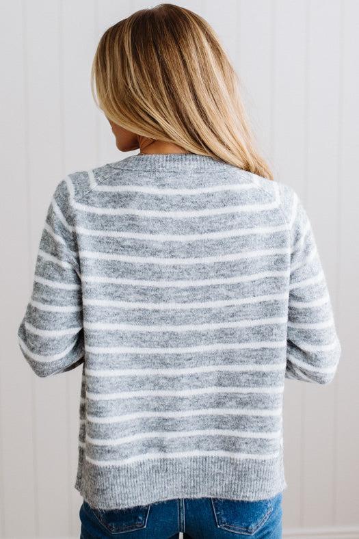 BLONDE WOMAN IN A BRIGHTLY LIT STUDIO WEARING A CREAM TANK AND DARK JEANS SHOWING A OUTSEAMED, LONG SLEEVE, FUZZY, CREW NECK STRIPED BUTTON DOWN SWEATER CARDIGAN IN HEATHER GREY.