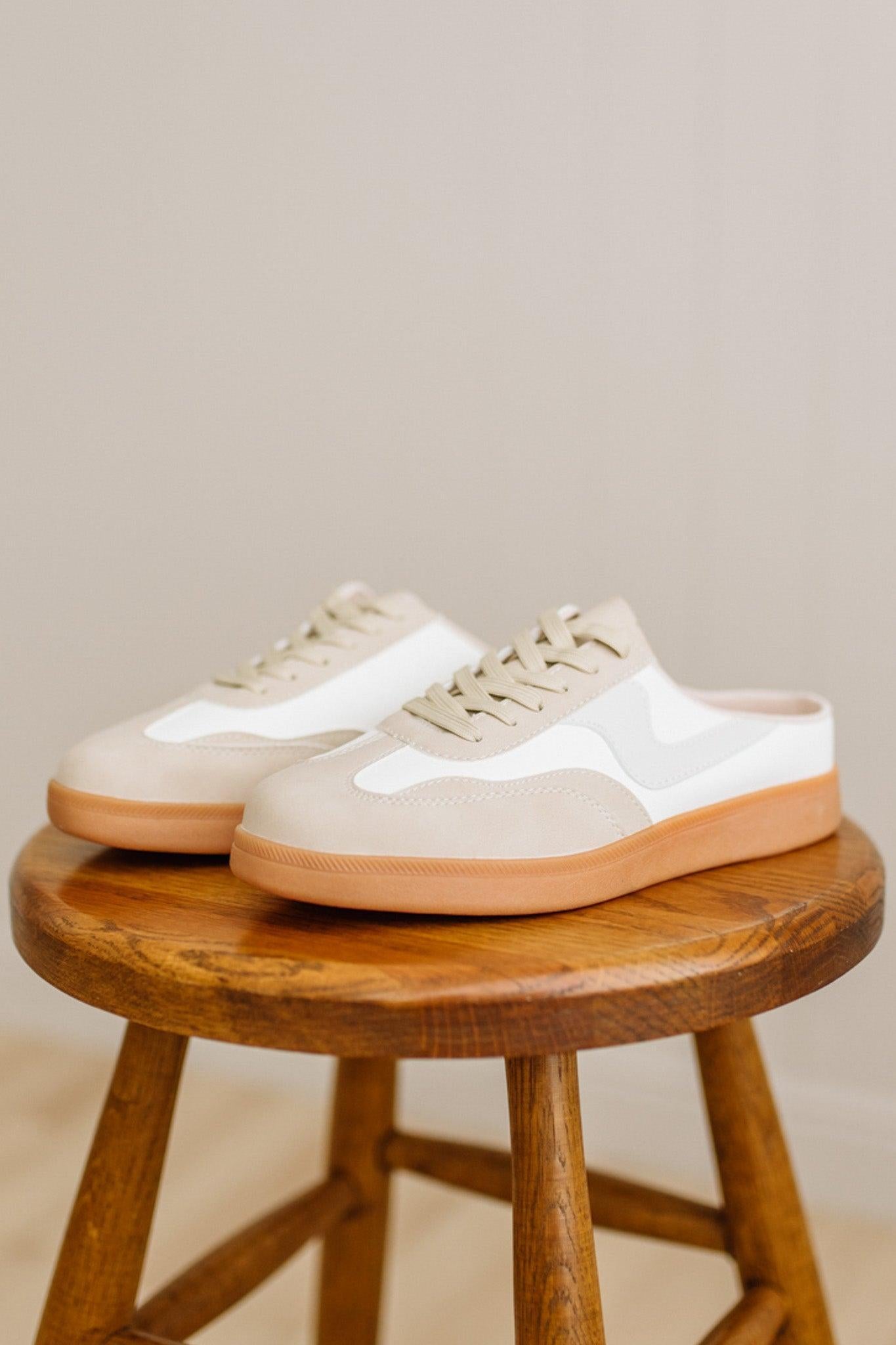 Pair of white sneakers with brown soles on a wooden stool against a beige background