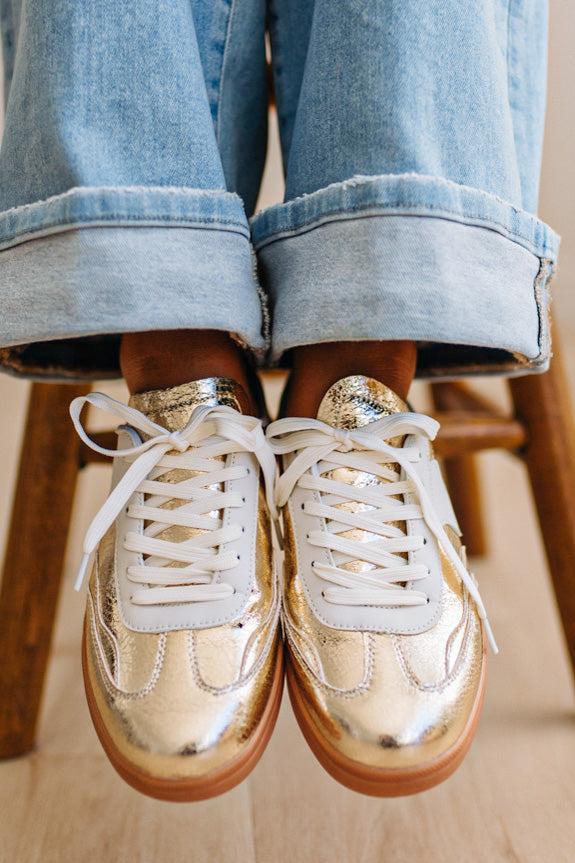 Woman wearing metallic gold and white slip-on sneakers featuring a bold swoosh pattern on the side, giving them a dynamic, sporty look. The durable gum sole provides excellent traction and a retro touch.
