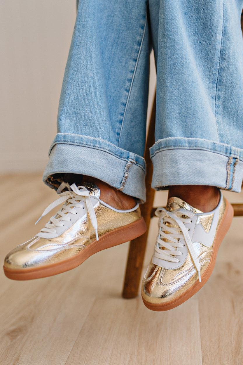 Woman wearing metallic gold and white slip-on sneakers featuring a bold swoosh pattern on the side, giving them a dynamic, sporty look. The durable gum sole provides excellent traction and a retro touch.
