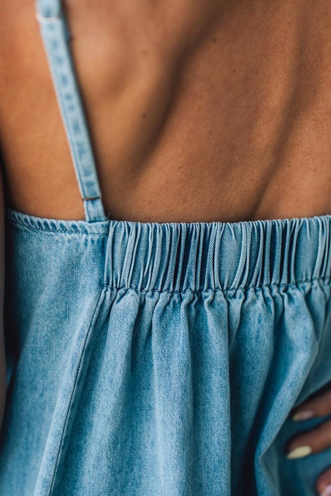 Blonde woman wearing a long denim maxi dress with adjustable spaghetti straps, side pockets, and a side slit.