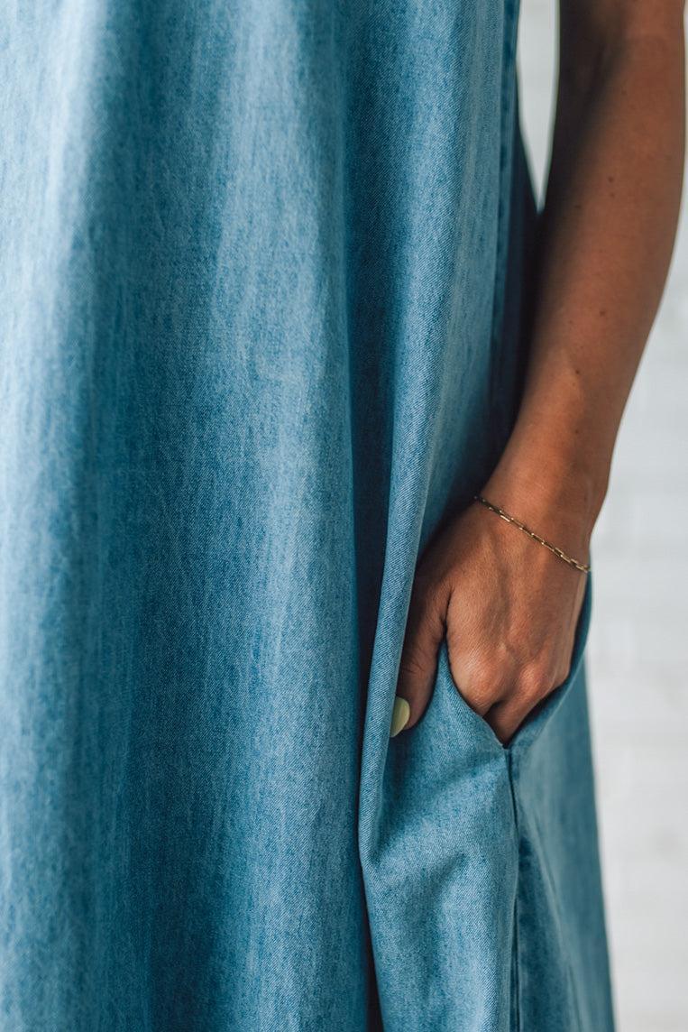 Blonde woman wearing a long denim maxi dress with adjustable spaghetti straps, side pockets, and a side slit.