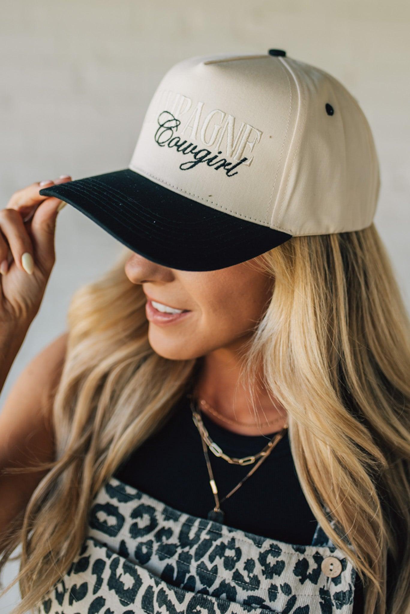 Blonde woman wearing a cream and black trucker hat with "Champagne Cowgirl" embroidered at the front.