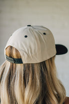 Blonde woman wearing a cream and black trucker hat with "Champagne Cowgirl" embroidered at the front.