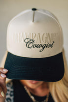 Blonde woman wearing a cream and black trucker hat with "Champagne Cowgirl" embroidered at the front.