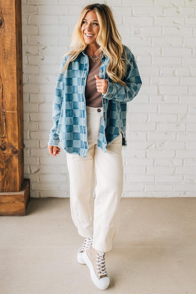Woman wearing a blue plaid shirt, brown top, white pants, and white sneakers in a room with a wooden floor and white brick wall.