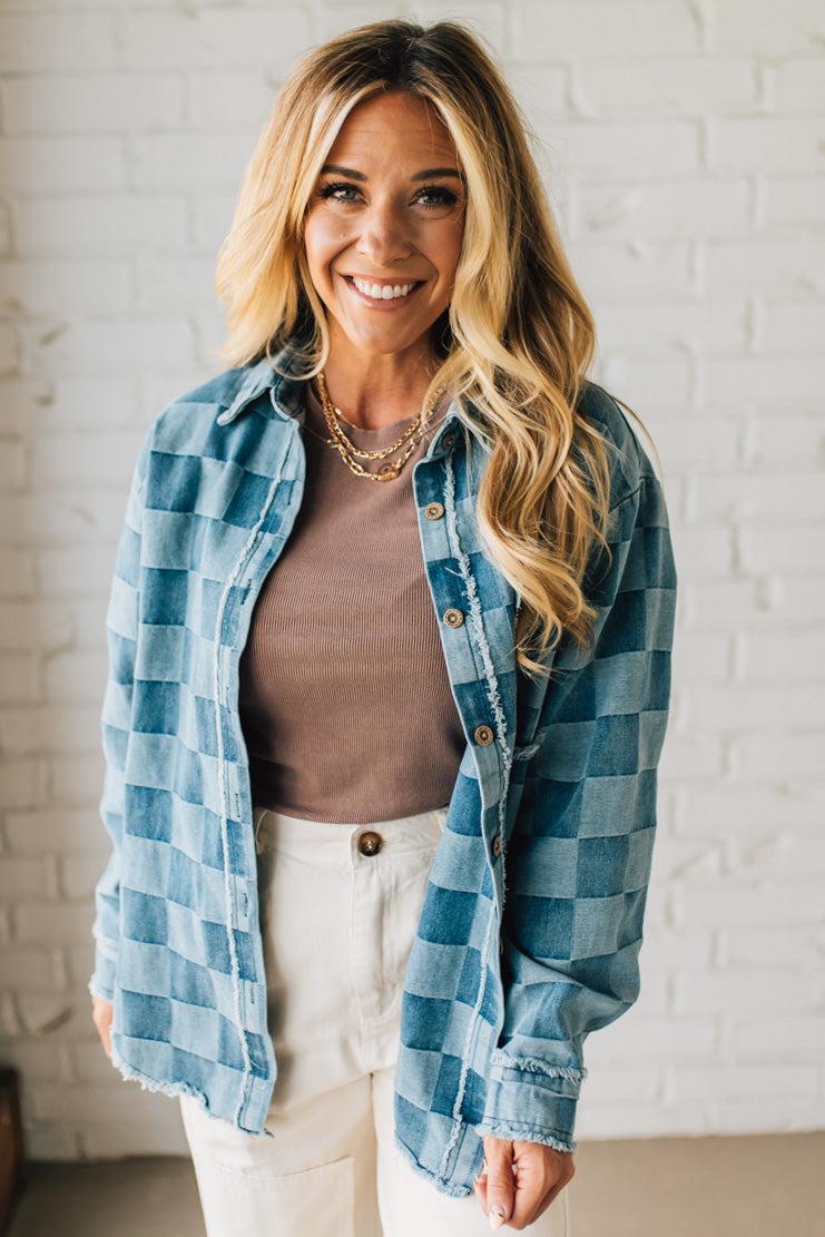 Woman wearing a blue plaid shirt over a brown top and white pants against a white brick wall.